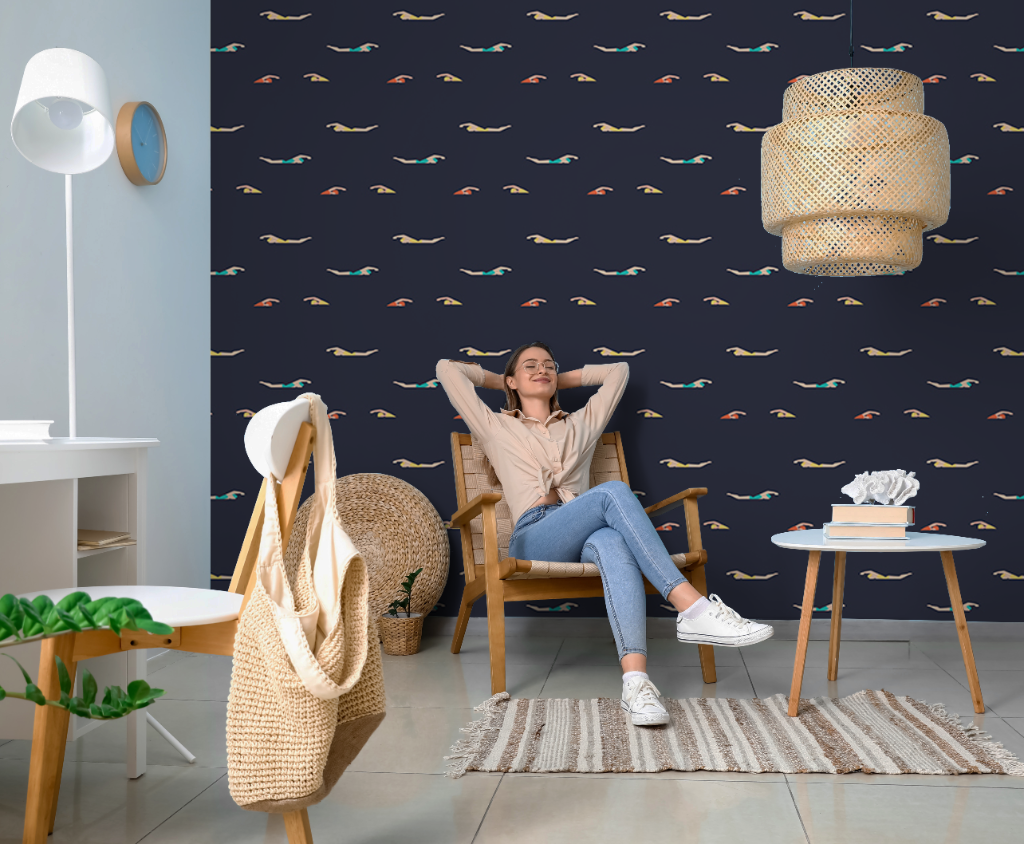 A woman relaxes in a rattan chair with her hands behind her head in a stylish room with Sixties Swimmers Wallpaper Mural, a white desk, woven accessories, and a hanging rattan lamp.