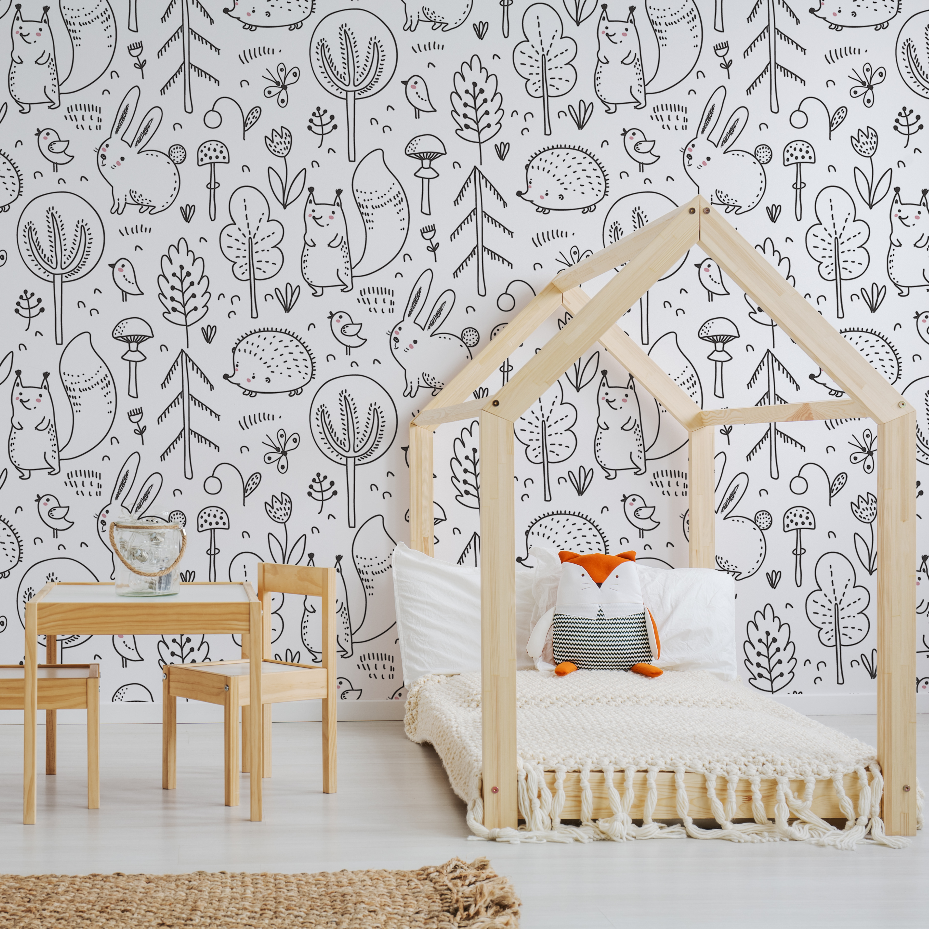 A bright children's room featuring a house-shaped wooden bed frame with a white mattress, decorated with an orange cushion, beside a small wooden table and chairs set against a Sketchbook Garden Wallpaper Mural by Decor2Go Wallpaper Mural.