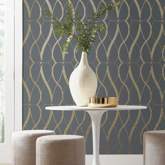 In a modern interior, a white vase with greenery sits on a round table against York Wallcoverings' Smoke & Gold Graceful Geo Wallpaper. Two light-colored stools and a small gold bowl from the Modern Metals Collection add an elegant touch.