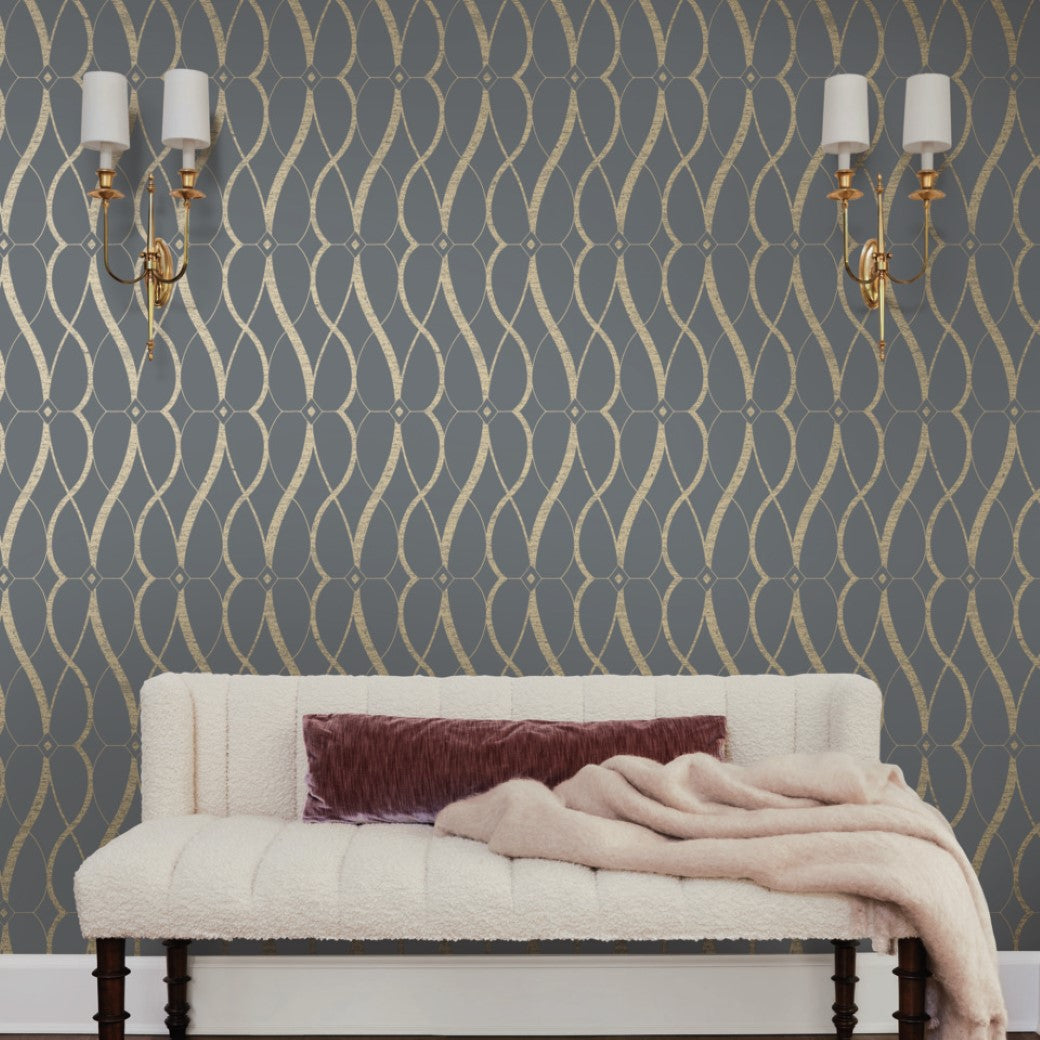 A plush white bench with a mauve cushion and beige throw sits against York Wallcoverings' Smoke & Gold Graceful Geo Wallpaper. Two elegant gold wall sconces with white shades hang above, adding sophistication enhanced by the smoke grey and gold hues from the Modern Metals Collection.