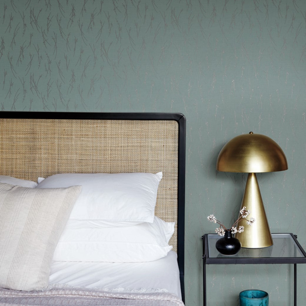 A modern bedroom showcases a rattan headboard, white pillows, and a neutral bedspread. A black side table from our Modern Metals Collection holds a gold lamp and decor. Smokey Blue & Silver Sprigs Wallpaper by York Wallcoverings features a leafy pattern in soft green.