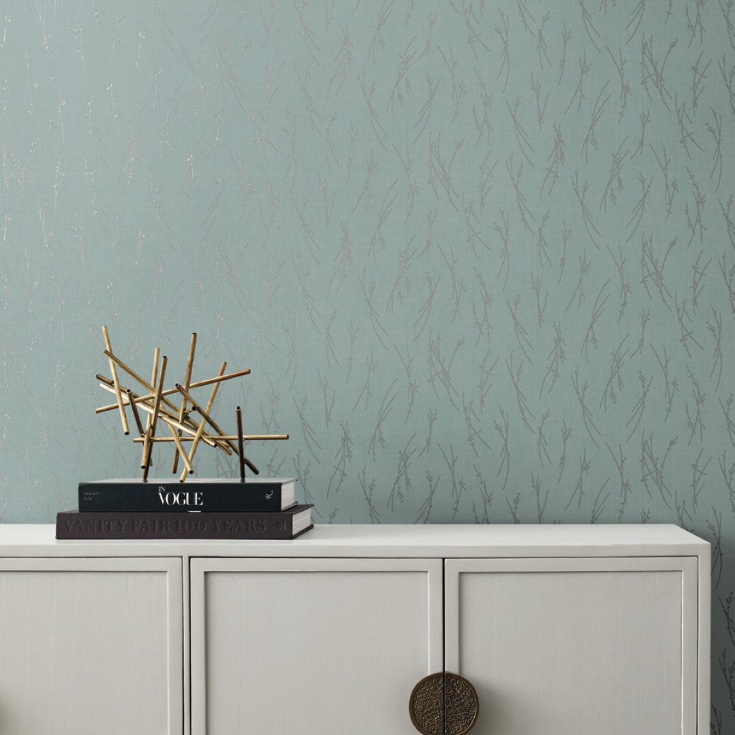 A minimalist room showcases a light grey cabinet with round handles from the Modern Metals Collection. Stacked books and a decorative gold sculpture sit on top, while York Wallcoverings' Smokey Blue & Silver Sprigs Wallpaper beautifully complements the modern look.