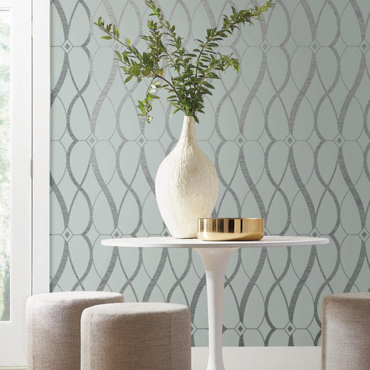 A white vase with green branches sits on a round white table paired with a gold bowl from the Modern Metals Collection. Two beige stools surround the setup. The background features York Wallcoverings' Spa & Silver Graceful Geo Wallpaper, with natural light streaming through a window on the left.