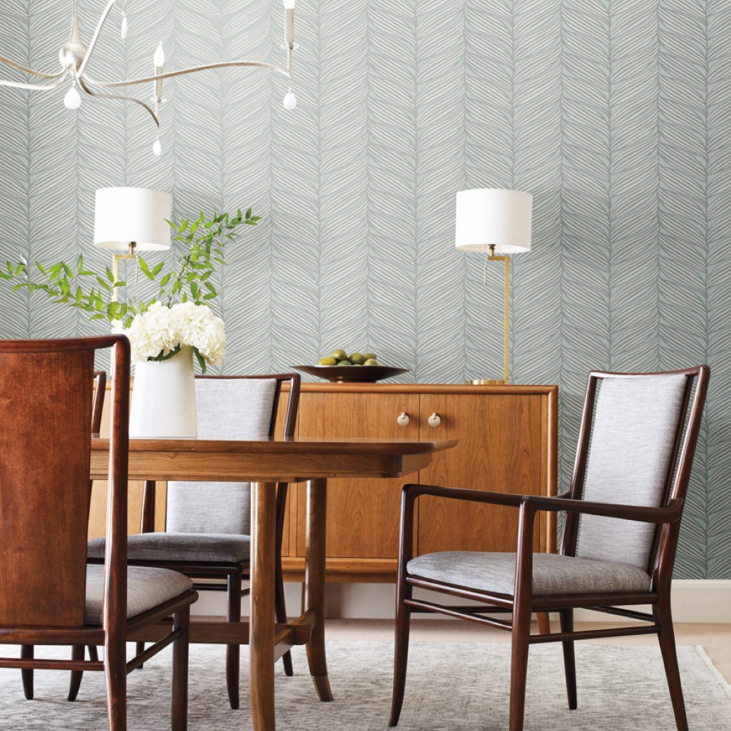Dining Room - A with a wooden table and chairs, a modern light fixture from the Modern Metals series, and a mid-century sideboard. York Wallcoverings' Spa & Silver Luminous Leaves wallpaper decorates the wall. Two lamps and a vase with palm leaves sit on the sideboard, while a rug covers the floor