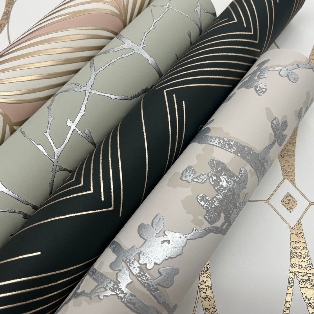 Three rolls of York Wallcoverings' decorative wallpaper are displayed in a fan arrangement, featuring gold geometric lines on dark green, silver floral on beige, and metallic palm motifs. One roll is the Blush & Gold Luminous Leaves Wallpaper from the Modern Metals Collection.