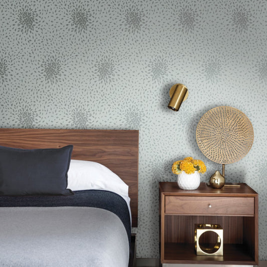 A stylish bedroom includes a wooden headboard bed with gray-black bedding, a wooden nightstand with yellow flowers and a lamp. The walls are adorned with York Wallcoverings' Spa & Silver Petite Leaves Wallpaper featuring subtle floral designs, complemented by a gold wall lamp.