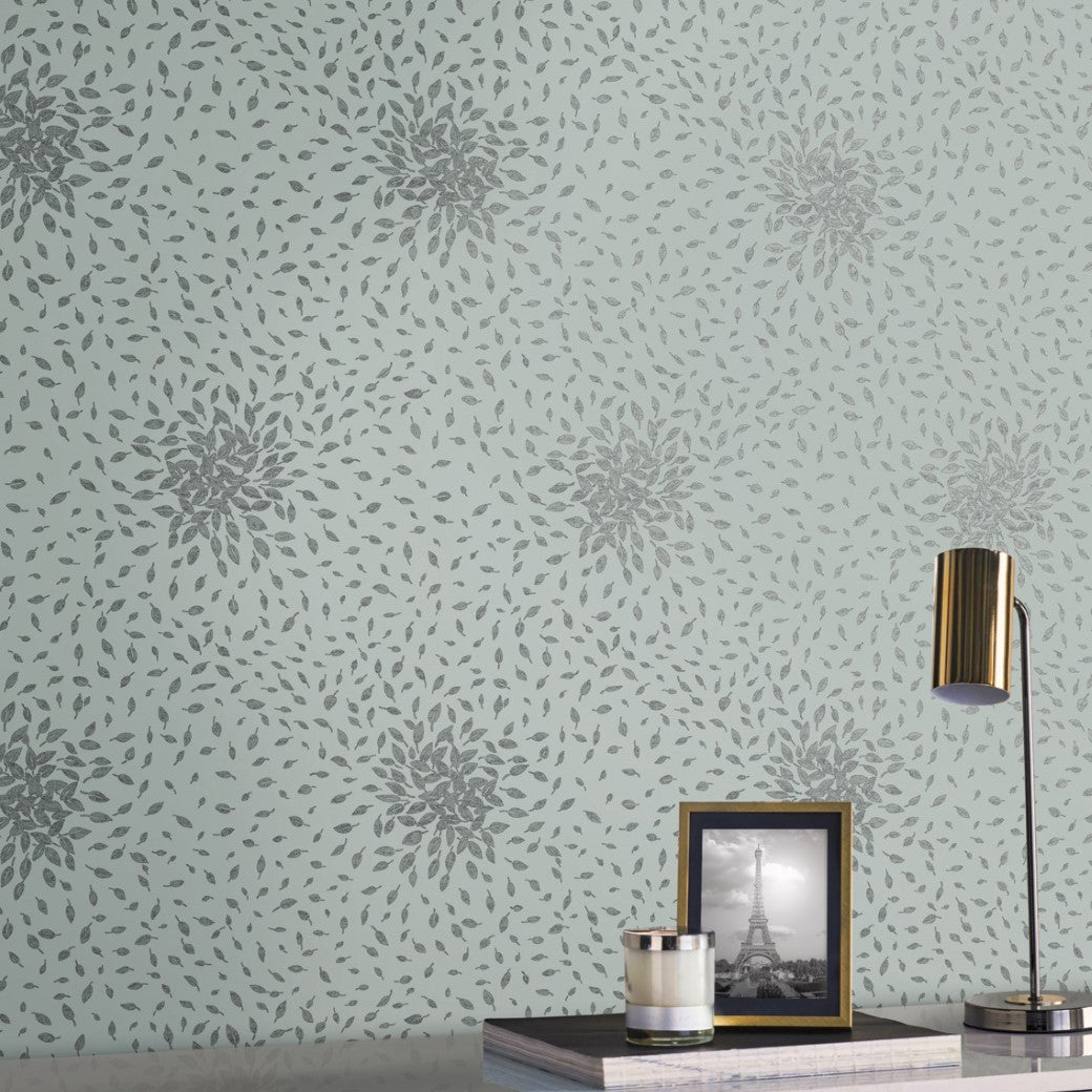 A decorative wall with Spa & Silver Petite Leaves Wallpaper by York Wallcoverings in green and gray. A table below holds a framed Eiffel Tower photo, a lit gold lamp, and a small silver cylinder from the Modern Metals Second Edition collection.