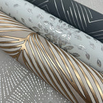 Four rolls of decorative wallpaper include thin silver lines, the Spa & Silver Petite Leaves Wallpaper by York Wallcoverings with silver leaves, metallic shapes from Modern Metals Second Edition, and gold linear patterns on beige.