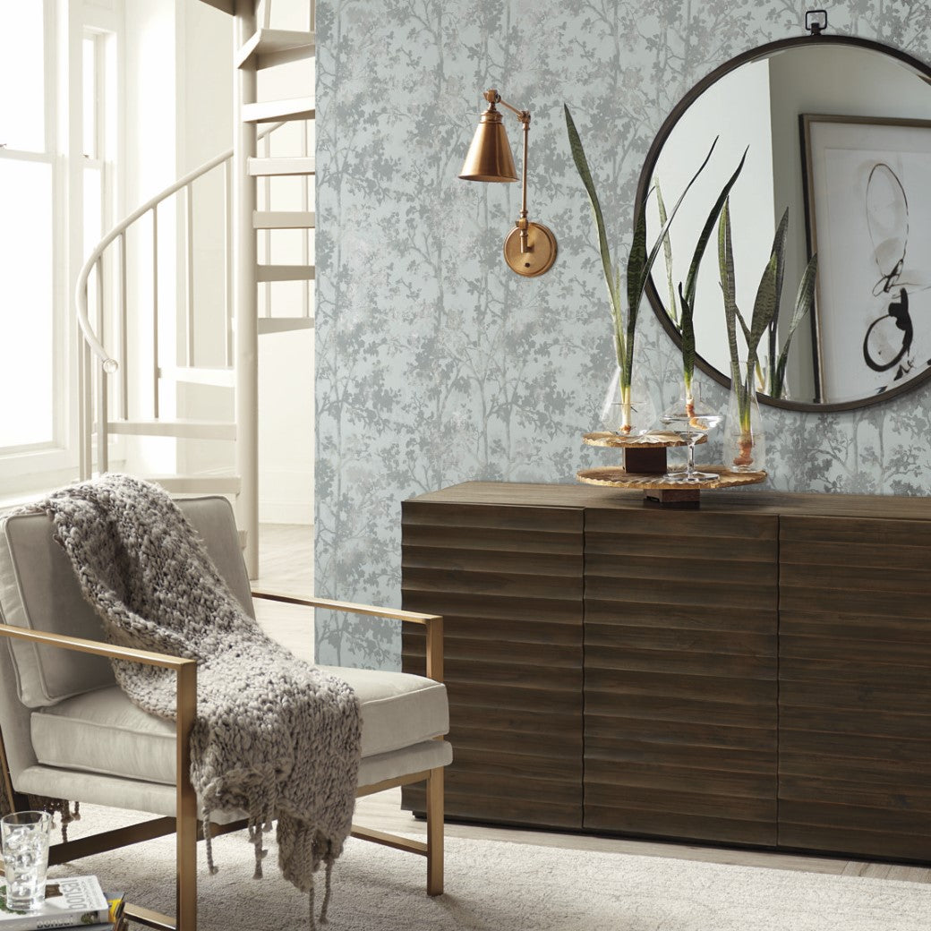 A cozy living room features a silver metallic armchair with a knitted throw. A wooden sideboard rests against York Wallcoverings' Spa & Silver Shimmering Foliage Wallpaper, topped with decor and a round mirror. A copper sconce lights the space, while a spiral staircase is nearby.