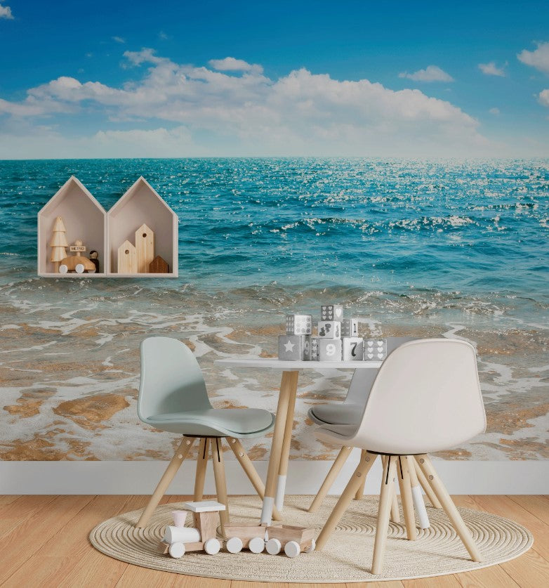 A small wooden table with two chairs, one light blue and one white, sits in a room adorned with a Summer Beach Waves Wallpaper Mural by Decor2Go Wallpaper Mural. A wall shelf shaped like house silhouettes holds small decorative items above the table, transforming the decor. A round rug lies on the floor.