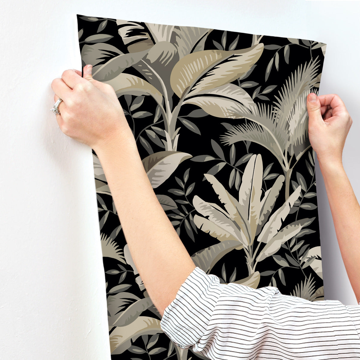 A person with a striped shirt is hanging the *Summerhouse Midnight Wallpaper Black, Brown (60 Sq.Ft.)* by *York Wallcoverings*, featuring a black background adorned with beige and gray leaves. The hands are holding the wallpaper straight against a white wall.