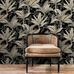 A wicker armchair with a cushioned seat and back rests on a wooden floor. The background features Summerhouse Midnight Wallpaper Black, Brown (60 Sq.Ft.) by York Wallcoverings, adorned with large, light-colored leaves, adding a serene touch to the space.