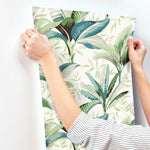 A person wearing a striped shirt is using both hands to hang a piece of Summerhouse Peacock Wallpaper Blue, Green (60 Sq.Ft.) by York Wallcoverings on a white wall. The person's left hand, adorned with rings, adds an element of elegance to the task, enhancing the tropical living vibe.