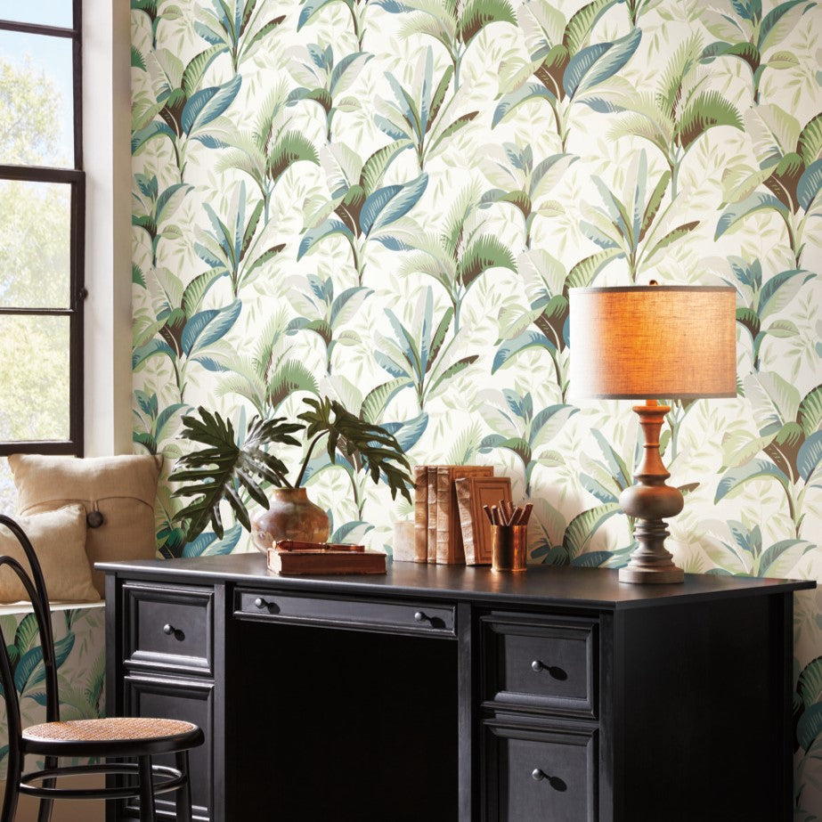 A home office setup featuring a black desk with drawers, a beige table lamp, books, and potted plants. The wall showcases York Wallcoverings Summerhouse Savanna Wallpaper Blue, Beige (60 Sq.Ft.) with easy installation. A cushioned window seat is visible on the left, next to a black chair with a circular backrest.