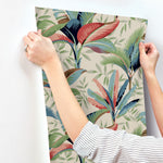 A person with a striped shirt and a ring is hanging up Summerhouse Midnight Wallpaper Black, Brown (60 Sq.Ft.) by York Wallcoverings that features a colorful, abstract tropical leaf design on a beige background. The Tropical wallpaper showcases leaves in shades of green, blue, red, and orange.