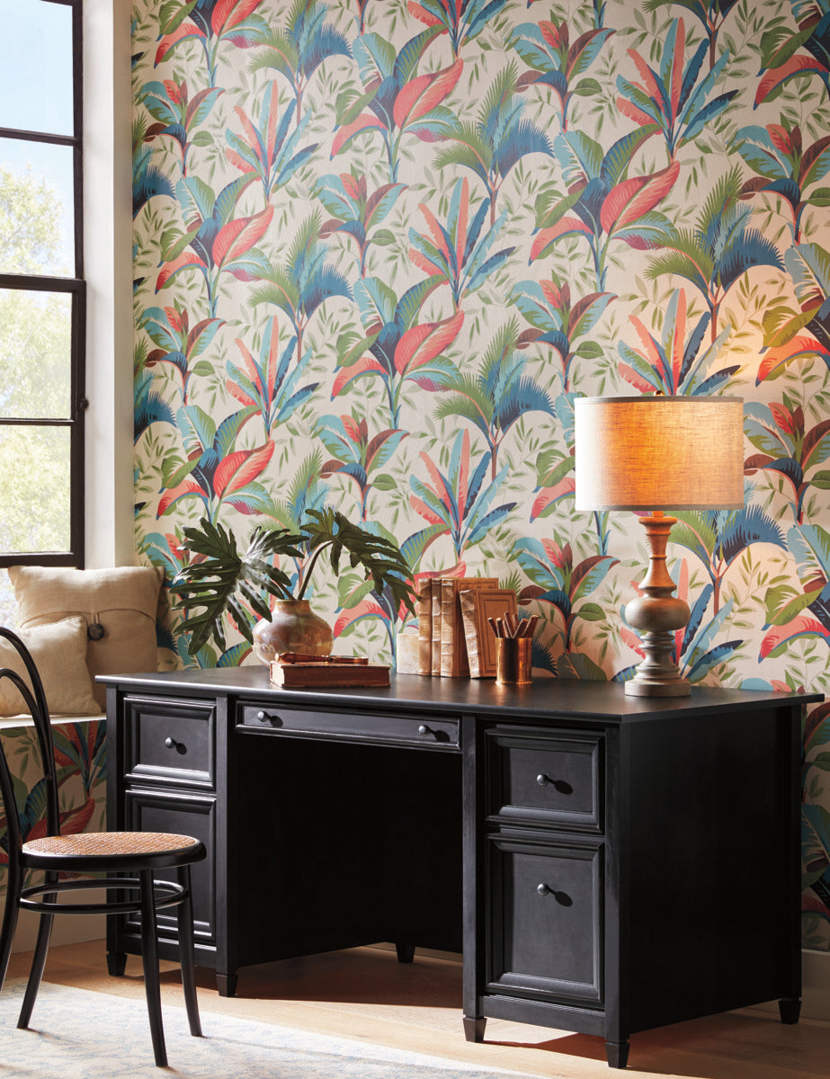 A home office with a black desk and chair against a vibrant, Summerhouse Savanna Wallpaper Blue, Beige (60 Sq.Ft.) by York Wallcoverings. The desk has two plants, a lamp, books, and office supplies. A window with natural light and a cushioned bench are on the left side. With its easy installation, this cozy ambiance is both inviting and practical.