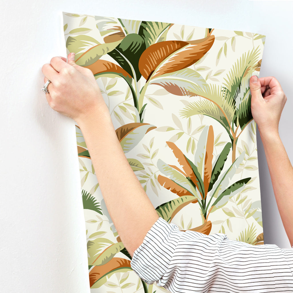 A person hanging a large, Summerhouse Midnight Wallpaper Orange, Green (60 Sq.Ft.) removable wallpaper on a white wall. Only their arms and hands are visible.