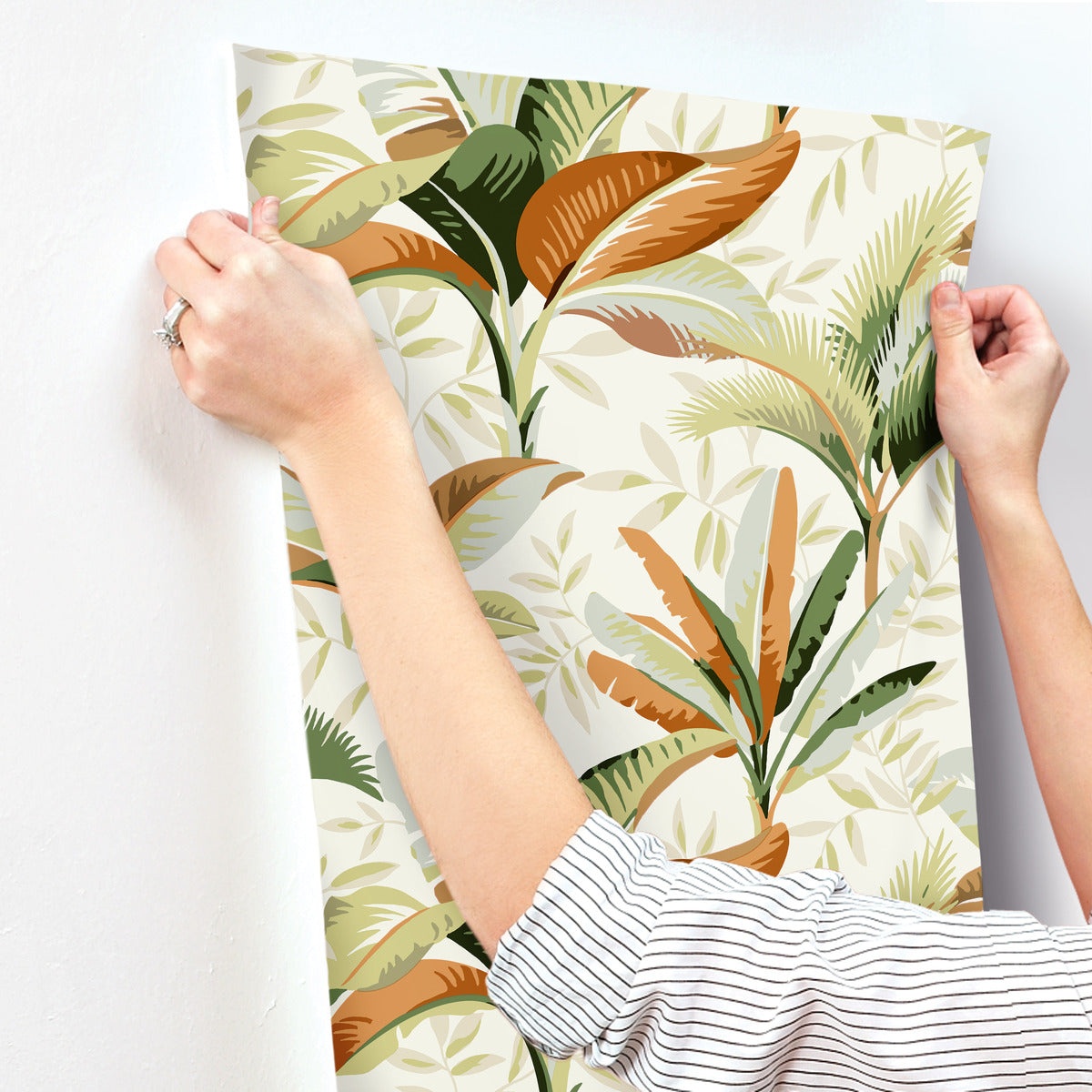 A person wearing a striped shirt is hanging removable York Wallcoverings Summerhouse Peacock Wallpaper Blue, Green (60 Sq.Ft.) with a tropical plant pattern on a white wall. The high-performance substrate design features various shades of green and brown leaves. The person's hands are visible, holding the edges of the wallpaper.