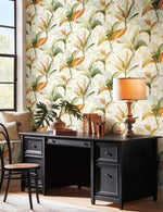 A home office features a large black desk against York Wallcoverings' Summerhouse Peacock Wallpaper Blue, Green (60 Sq.Ft.) with green and yellow leaf patterns. On the desk are books, a lamp, potted plants, a decorative rattan tray, and a framed photo. A wooden chair with a woven seat is in front of the desk, creating a serene tropical living vibe.