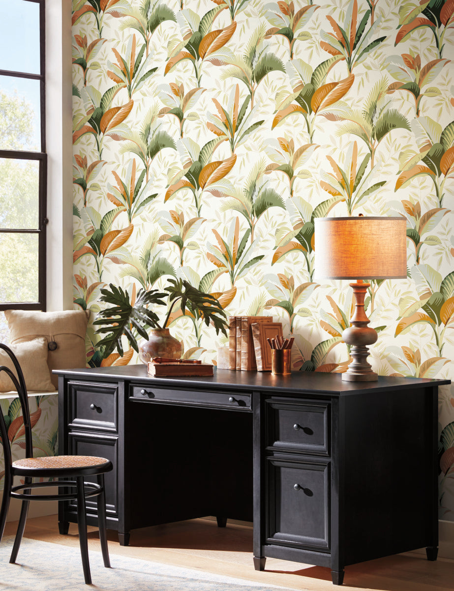 A home office with a black desk, a chair, and various office supplies. A beige lamp with an orange shade is on the desk alongside books and a plant. The wall behind features easy removable Summerhouse Midnight Wallpaper Black, Brown (60 Sq.Ft.) by York Wallcoverings adorned with green, yellow, and brown leaves. Sunlight streams through large windows.