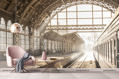 A conceptual design of a vintage railway station transformed into a stylish living space features a modern pink chair, sleek floor lamp, and elegant Decor2Go Wallpaper Mural under a grand arched roof with sunlit tracks.