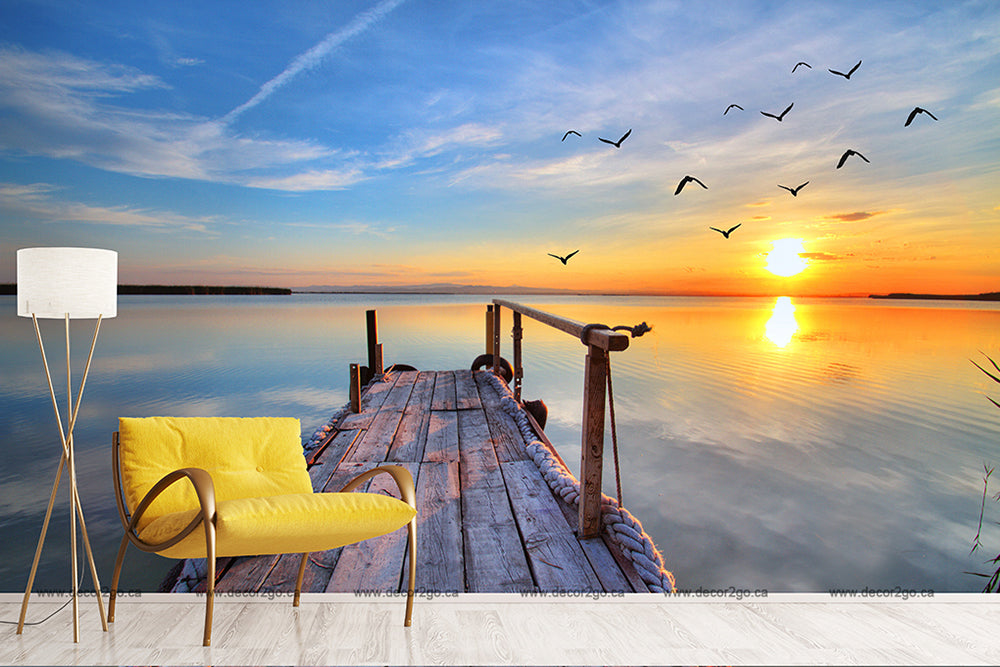 An interior view blending into an outdoor scene at a lakeside retreat with a yellow chair and white floor lamp on a wooden floor, extending to a dock over a tranquil lake with Decor2Go Wallpaper Mural's Sunset Birds Flying Wallpaper Mural.