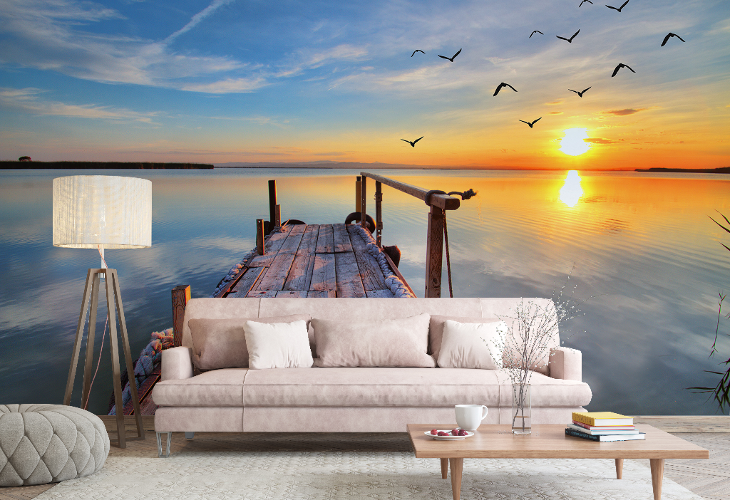 A surreal lakeside retreat setup on a wooden dock featuring the Decor2Go Wallpaper Mural "Sunset Birds Flying" on a comfortable sofa, coffee table, lamp, and scattered cushions, overlooking a tranquil lake with flying birds.