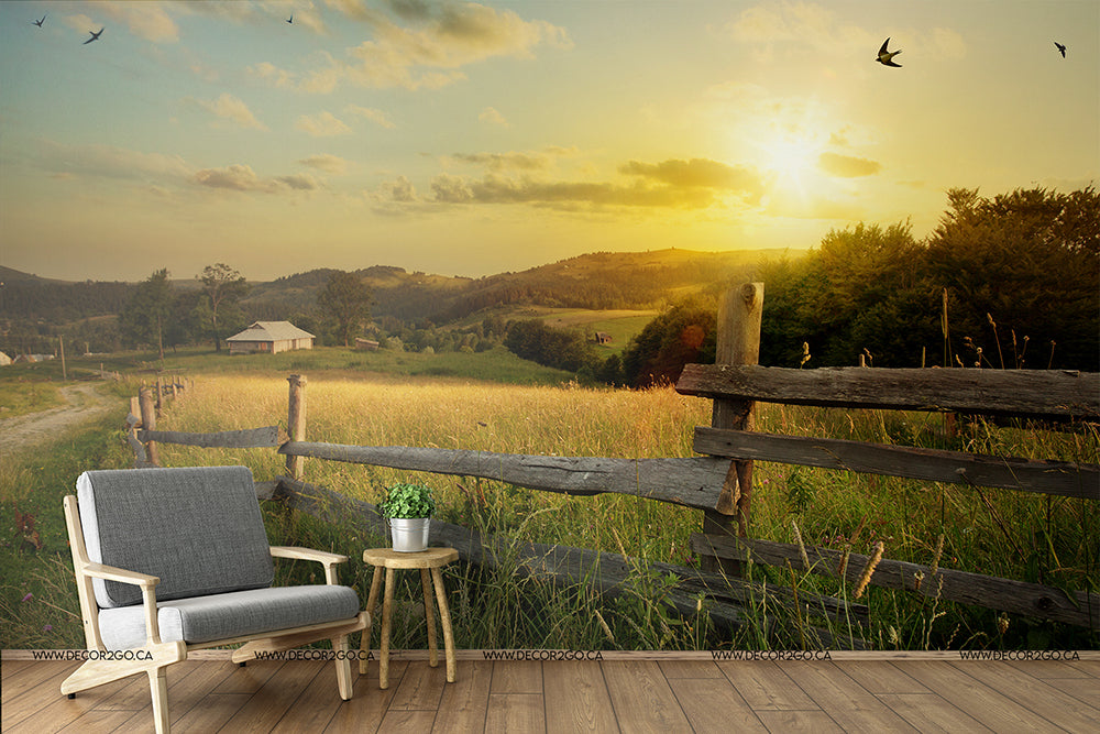 A serene rural landscape at sunset, featuring a vintage armchair and table beside a wooden fence, overlooking a field with a farmhouse in the distance and birds flying in the golden sky is beautifully captured in the Sunset Farm Wallpaper Mural by Decor2Go Wallpaper Mural.