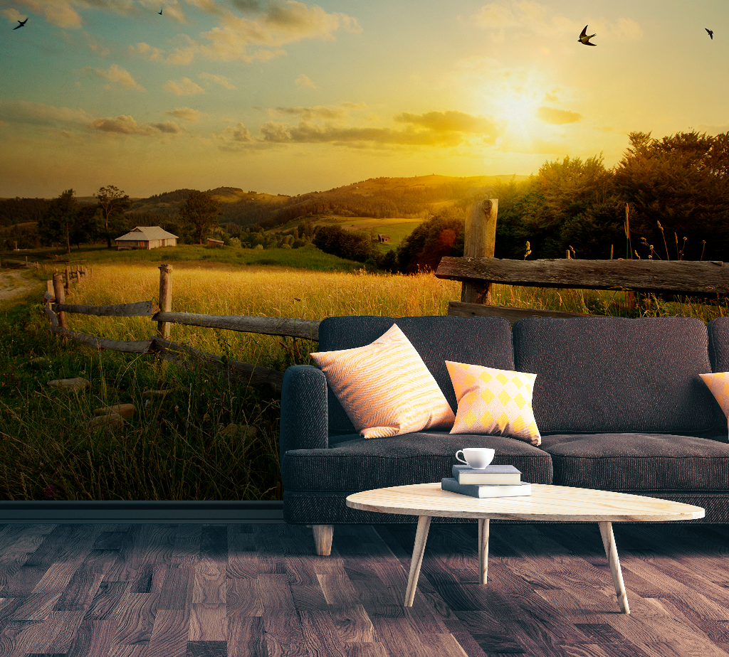 A surreal image blending an indoor scene with an outdoor landscape; a gray sofa and a coffee table inside a room that seamlessly transforms into Decor2Go Wallpaper Mural featuring a rural sunset scene with a house and trees