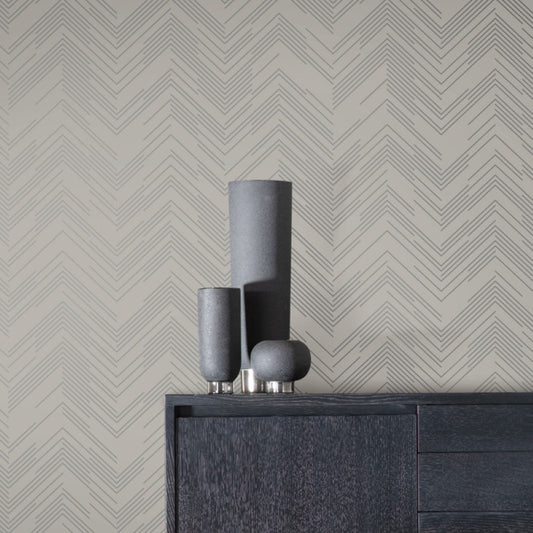 Vases in different sizes rest on a dark wood cabinet, beautifully contrasted by York Wallcoverings' Taupe & Silver Polished Chevron Wallpaper, capturing Geometric Elegance.