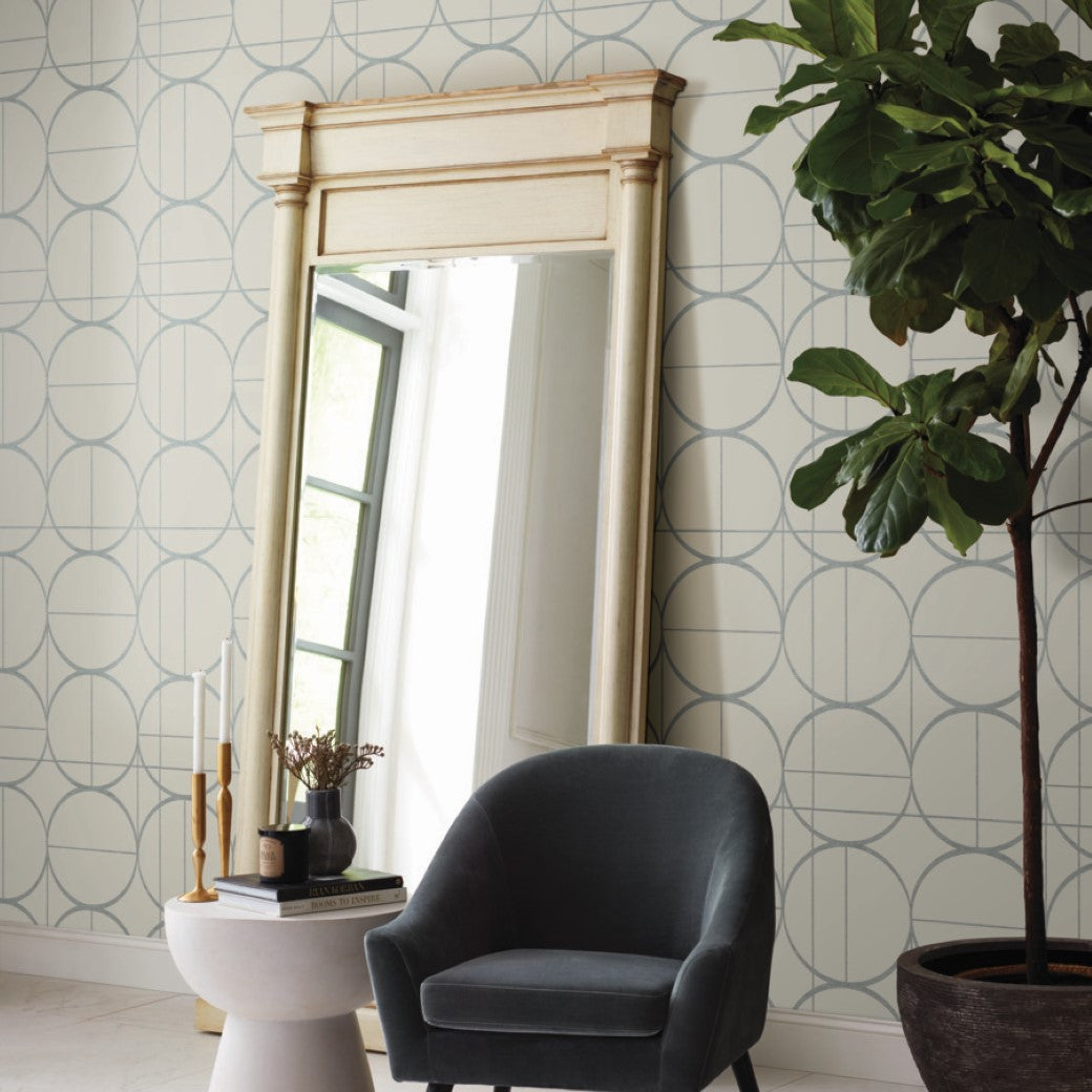 A stylish room features a tall, wooden-framed mirror reflecting light from a window. Nearby is a dark gray upholstered chair with a small table adorned with candles and books. A large potted plant adds greenery against York Wallcoverings' Taupe & Silver Sun Circles Wallpaper's geometric pattern.