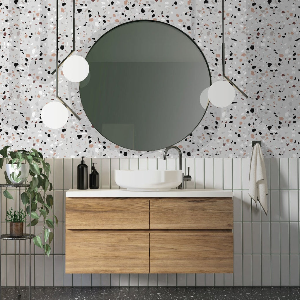A modern bathroom featuring a round mirror hanging above a wooden vanity with a white basin. The wall is adorned with Decor2Go Wallpaper Mural in luxurious terrazzo design. There are hanging spherical lamps and a potted plant to the left.