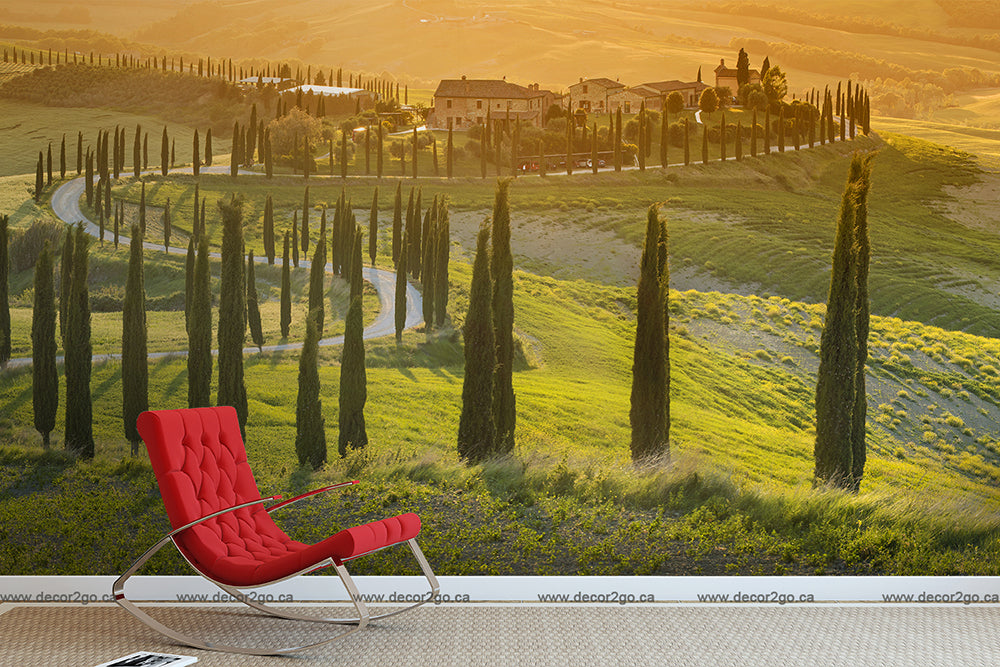 A red lounge chair faces a scenic view of vibrant green Tuscan hills, with cypress trees lining a winding road leading to a distant village, bathed in warm orange sunset light featuring the Toscana Trail Wallpaper Mural by Decor2Go Wallpaper Mural.