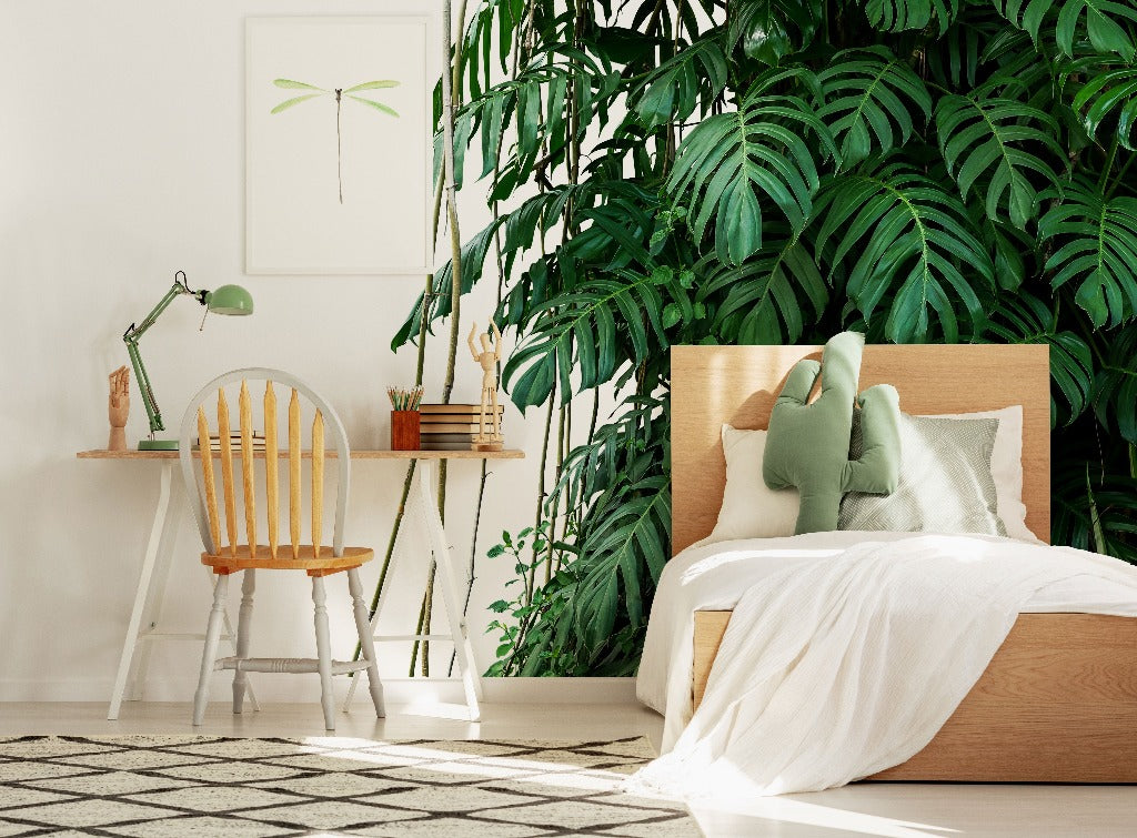 A bright and cozy bedroom corner with a large, leafy indoor plant, wooden bed and desk with a chair, and soothing green-themed Decor2Go Wallpaper Mural featuring the Tropical Jungle Leaves Wallpaper Mural.