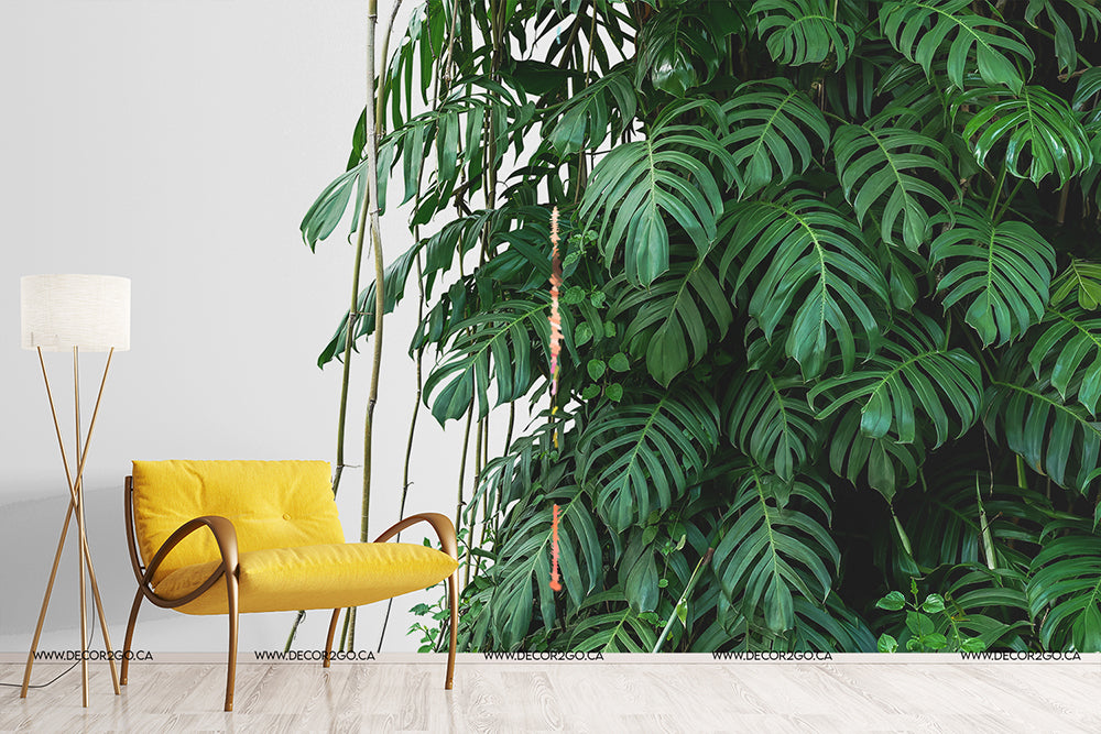 A modern living room corner with a large, lush green wall of Tropical Jungle Leaves Wallpaper Mural from Decor2Go Wallpaper Mural. A minimalist yellow armchair and a white floor lamp are to the left, enhancing the serene indoor garden vibe.