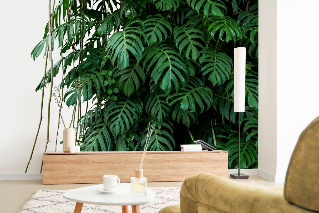 A cozy living room corner with a Tropical Jungle Leaves Wallpaper Mural from Decor2Go Wallpaper Mural in the background, a small wooden table with candles in the foreground, and part of a comfy chair visible.