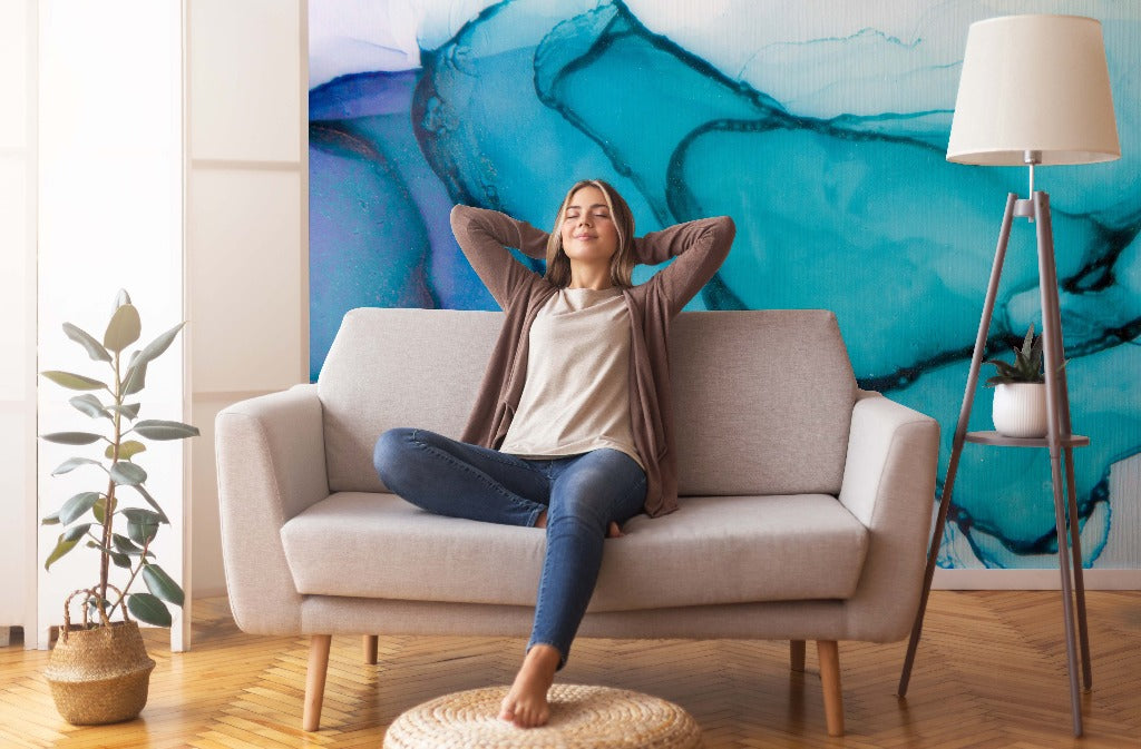 A woman relaxes on a sofa with her hands behind her head in a cozy living room, surrounded by plants and vibrant Blue Ocean Waves Wallpaper Mural from Decor2Go.