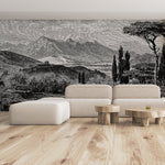 A contemporary living room with light wood flooring showcases a minimalist beige sofa paired with three wooden coffee tables. The wall is adorned with the "Tuscan Serenity Wallpaper Mural" by Decor2Go, featuring an intricate black-and-white depiction of the Tuscan countryside, complete with mountains, trees, and a rural scene.