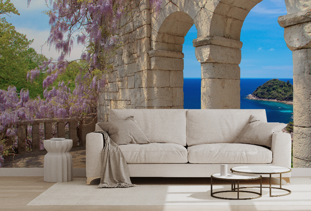 A serene living room setup with a white couch, rug, and small tables under stone arches featuring a Tuscany Veranda View Wallpaper Mural from Decor2Go Winnipeg overlooking a scenic ocean view and green island, accented by purple wisteria.