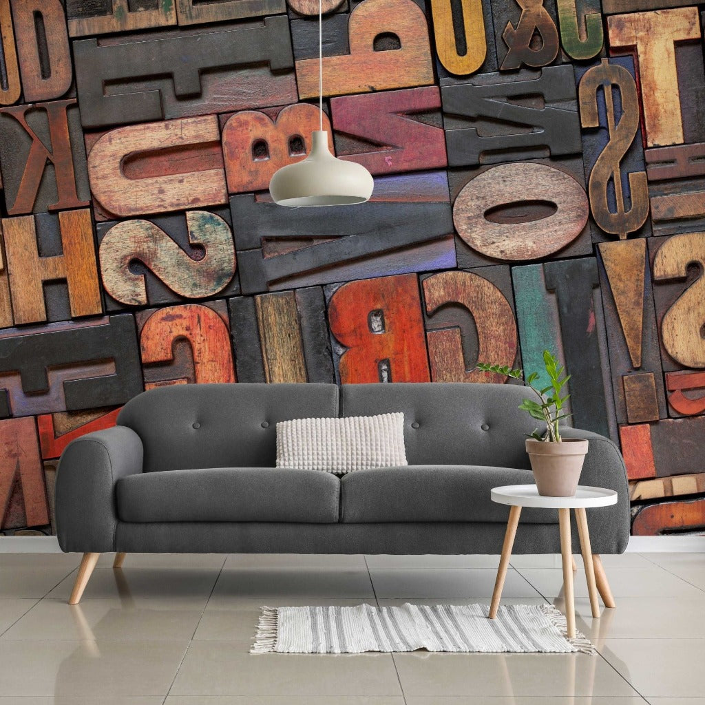 A stylish living room with a gray sofa, decorative cushions, a small white coffee table with a potted plant, a white rug, and a large colorful Decor2Go Wallpaper Mural with various letters.