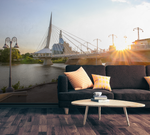 A stylish living room setup with a black couch, patterned cushions, and a white coffee table, featuring a Decor2Go Urban Bridge Wallpaper Mural and river during sunset.