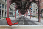A red modern chair positioned in the center of an empty street in Queens NYC, under an urban railway track. Light casts long shadows on the concrete, enhanced by a Decor2Go Wallpaper Mural nearby, giving a stark
