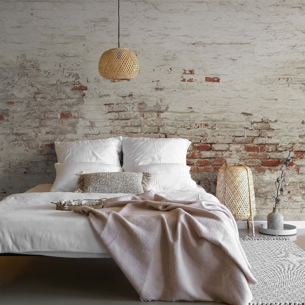 A cozy bedroom featuring an unmade bed with white bedding and a beige throw, against a rustic exposed brick wall enhanced by Decor2Go Wallpaper Mural. A wicker pendant light, a tall lantern, and simple