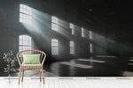 An empty industrial loft with large, architectural windows casting beams of natural sunlight across the floor, featuring a single rattan chair with a green cushion on the right foreground, and a Decor2Go Wallpaper Mural in Versailles design hanging on the back wall.