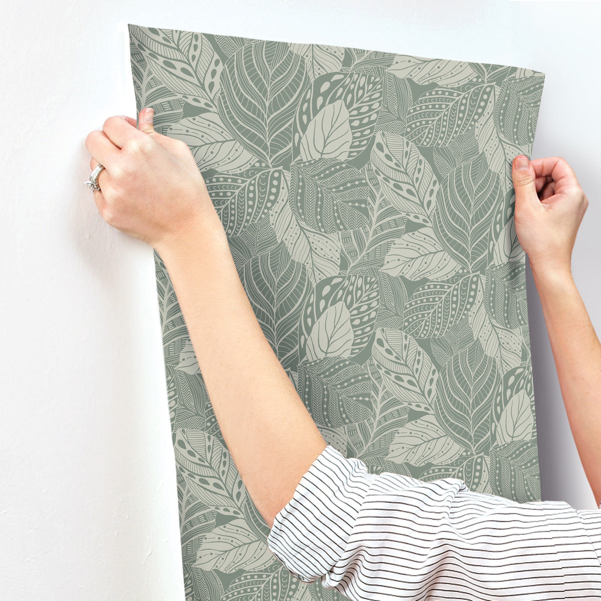 A person is hanging a sheet of York Wallcoverings Vinca Eucalyptus Wallpaper Green (60 Sq.Ft.) with a green and cream leaf pattern on a white wall. The individual, wearing a striped long-sleeve shirt and a ring on their left hand, effortlessly manages the durable non-woven material that is easy to hang and remove.