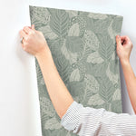 A person with rolled-up sleeves is applying York Wallcoverings Vinca Smokey Blue Wallpaper Blue (60 Sq.Ft.) to a white wall. The wallpaper, made of durable material, features intricate leaf designs in shades of green and beige. The person's hands are pressing the wallpaper against the wall to smooth it out.