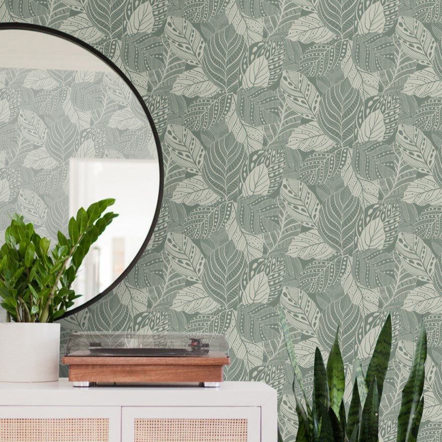 A stylish interior setup with a round mirror reflecting part of a room, a potted Zamioculcas plant on a white cabinet, a wooden turntable, and lush green leafy Vinca Glint Wallpaper Metallic (60 Sq.Ft.) by York Wallcoverings in the background, beautifully complementing the indoor plants.