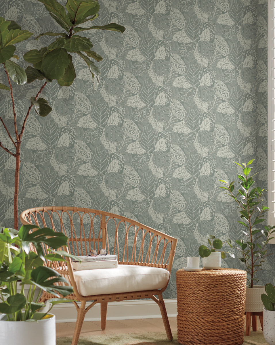 A cozy corner of a room featuring a rattan two-seater sofa with a white cushion, against a wall with Vinca Eucalyptus Wallpaper Green (60 Sq.Ft.) by York Wallcoverings. There are potted plants and a woven basket in the serene setting.