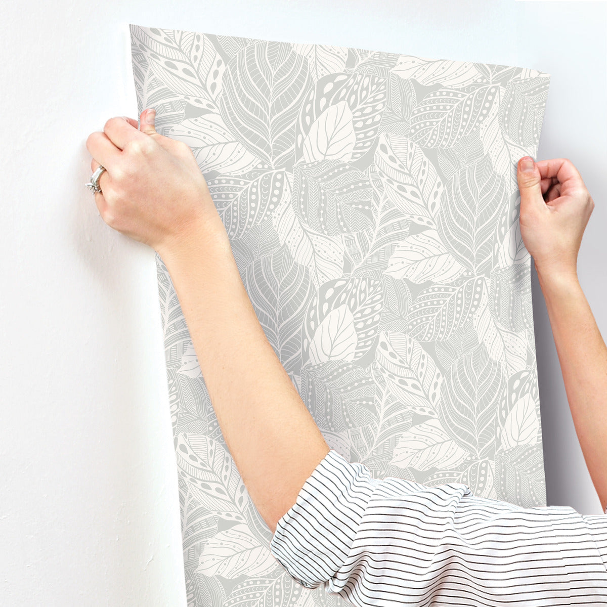 A person wearing a striped shirt is applying Vinca Glint Wallpaper Metallic (60 Sq.Ft.) by York Wallcoverings to a white wall. The wallpaper, made of durable non-woven material, features detailed, monochromatic leaves in various light and dark tones, creating a subtle and elegant design reminiscent of a textural canopy of vines.