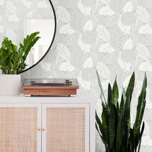 A stylish room featuring a white cabinet with a cane front, topped with a modern wooden turntable and a white potted plant. Beside the cabinet stands a tall leafy plant. A round mirror hangs on a wall adorned with York Wallcoverings Vinca Flint Wallpaper Grey (60 Sq.Ft.), showcasing its gray and white leaf-patterned design.
